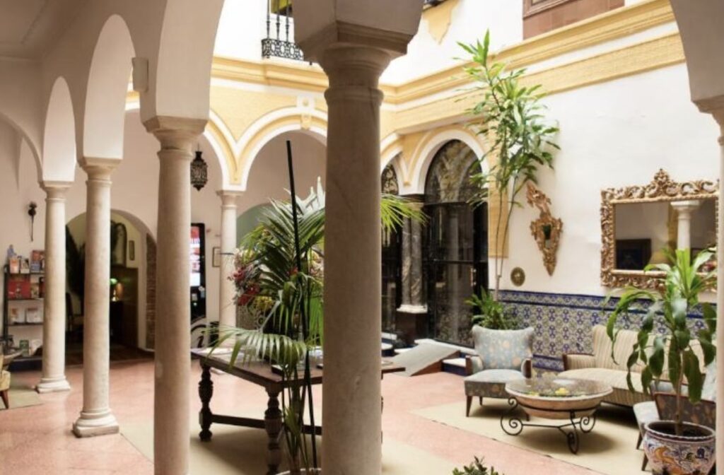 Columned Andalusian courtyard with white arches, blue-and-white tiled walls, leafy potted palms, a gilt mirror and traditional seating around a table