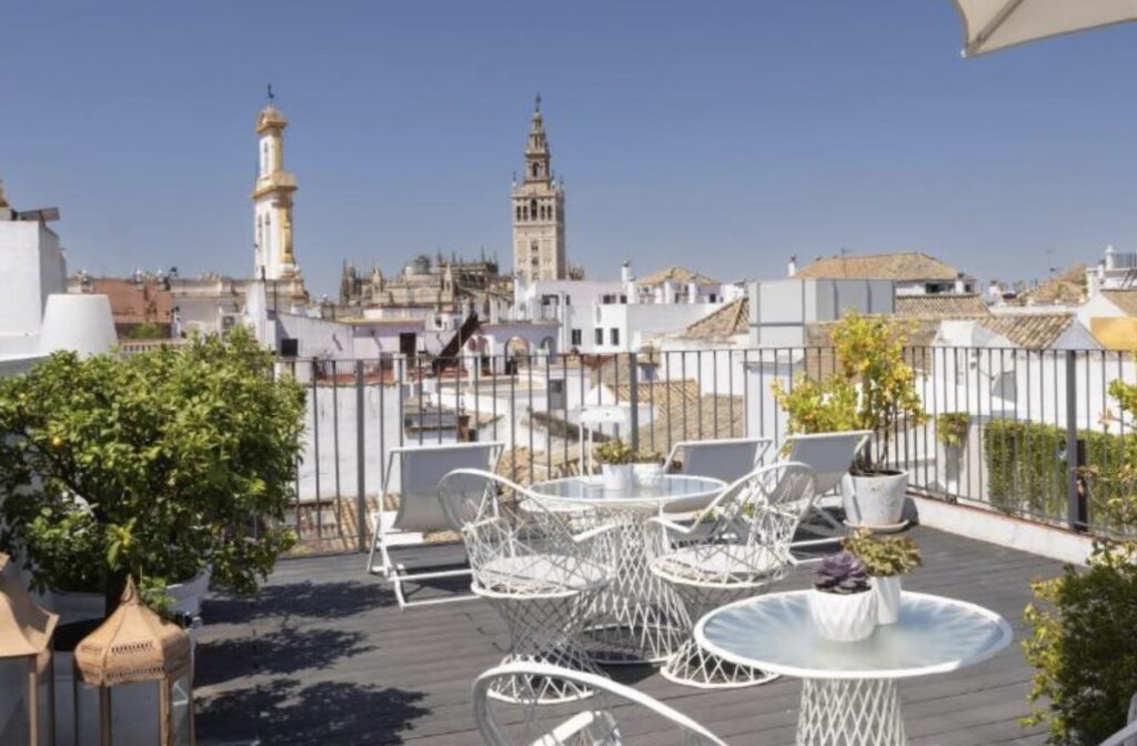 White-railed rooftop terrace with a lantern and drinks on a side table, roof tiles below and the tip of the Giralda catching evening light