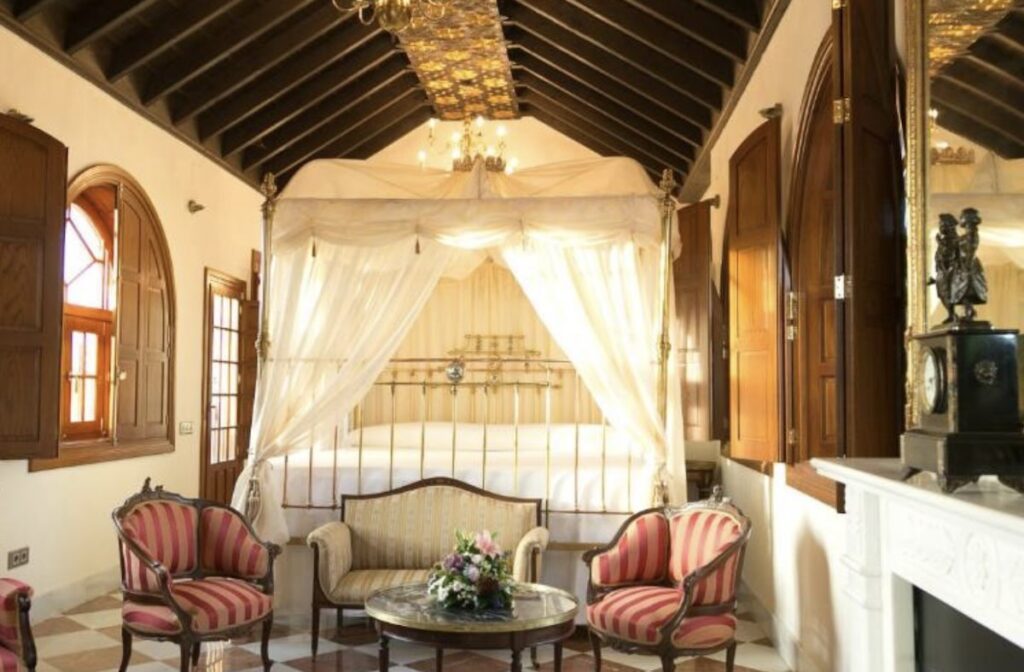 Ornate suite with a brass four-poster canopy bed, high beamed ceiling, arched wooden shutters, a marble fireplace and striped antique armchairs