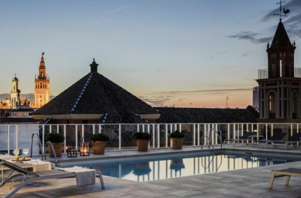 Rectangular rooftop pool ringed by loungers and potted shrubs at dusk, with Seville's illuminated Giralda bell tower on the skyline behind