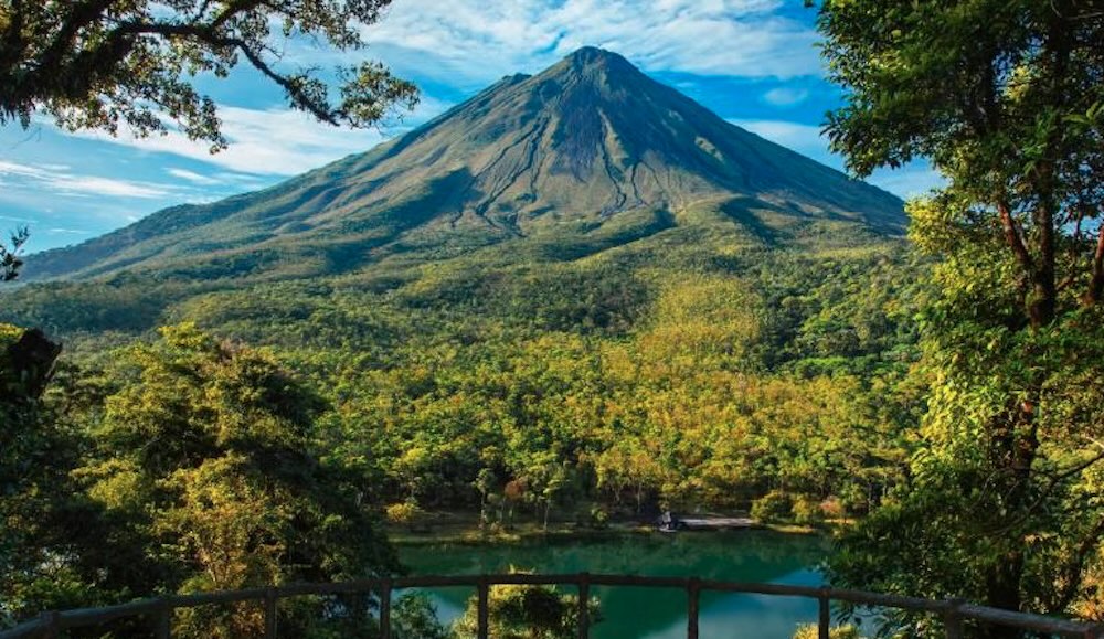 Arenal Volcano seen from a wooden viewing platform, its bare cone rising above rainforest and a green lake in the valley below