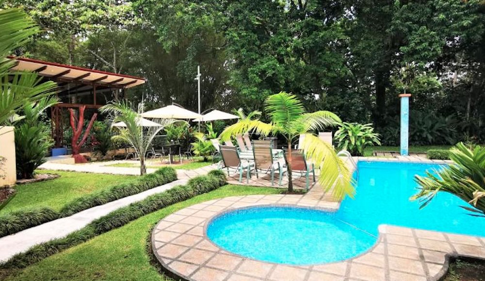 Small round paddling pool and a larger curved swimming pool set in a lawned garden with palms, parasols and a covered dining terrace