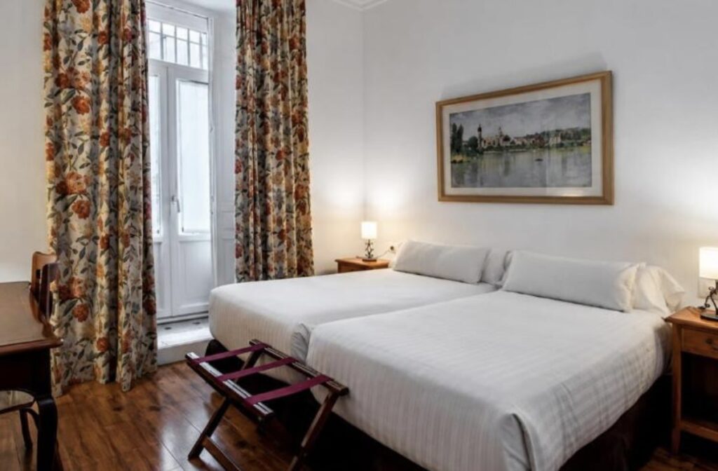 Twin beds with white linen in a traditional room, tall floral curtains at a balcony window, wooden floor and a framed river landscape painting