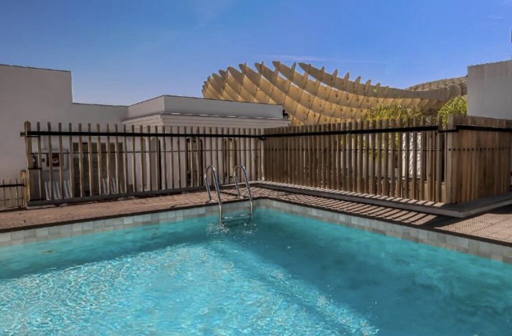 Turquoise rooftop pool in bright sun, with the curving timber lattice of Seville's Las Setas Metropol Parasol rising just behind the fence