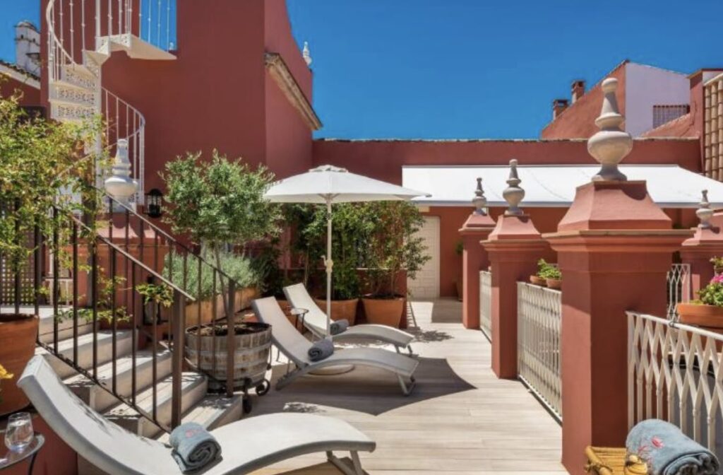 Rooftop sun terrace with grey loungers and rolled towels, potted olive trees and a spiral staircase against warm terracotta-red walls