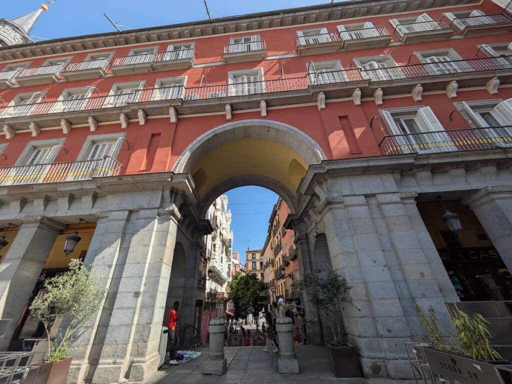 Arched stone gateway leading into Plaza Mayor in Madrid, framing a narrow street beneath the red-painted facade