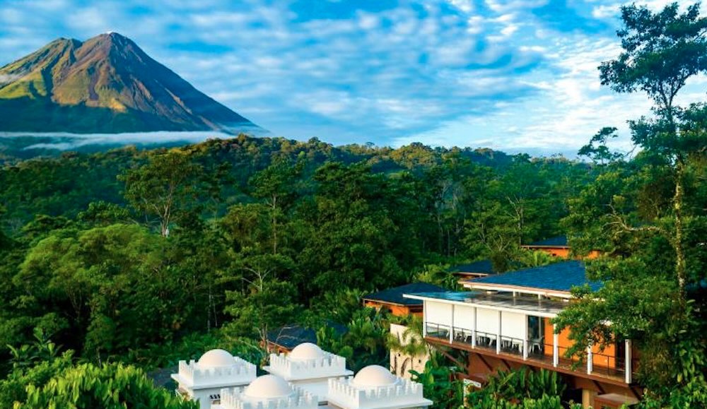 Arenal Volcano under scattered cloud, with the white domes and glass-fronted lodges of a hillside resort in the foreground