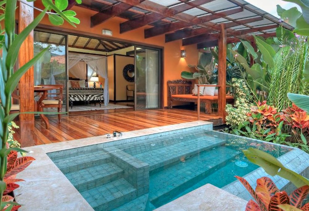 Private thermal plunge pool with steps, set into a timber deck outside a villa with a four-poster bed visible through the doors