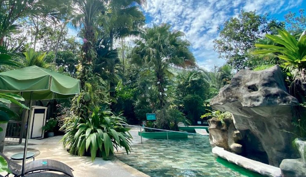 Pale green thermal pool winding past a large boulder and a green parasol, edged with palms and tropical planting under a blue sky