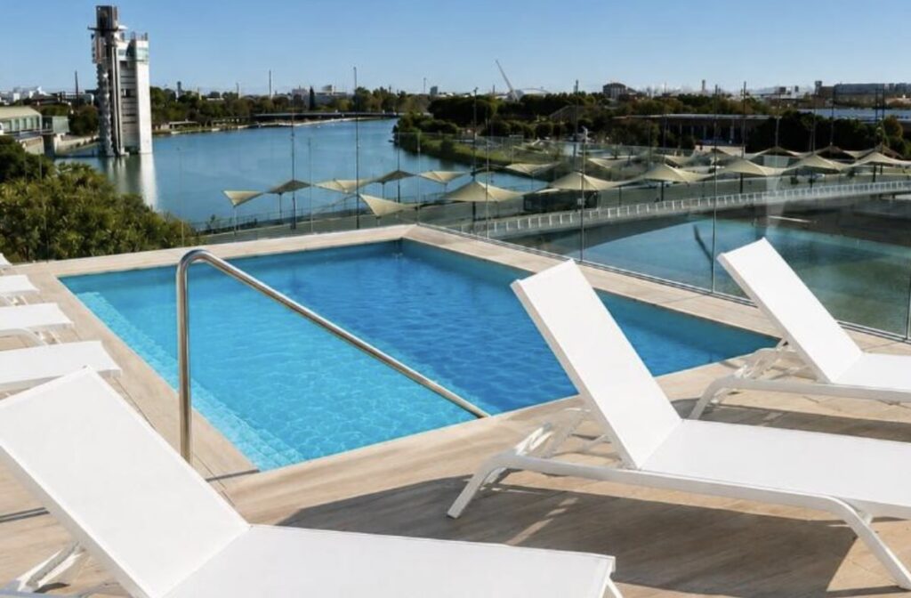 Small square rooftop plunge pool with white loungers beside a glass rail, looking across the Guadalquivir river to trees and a tower on the far bank