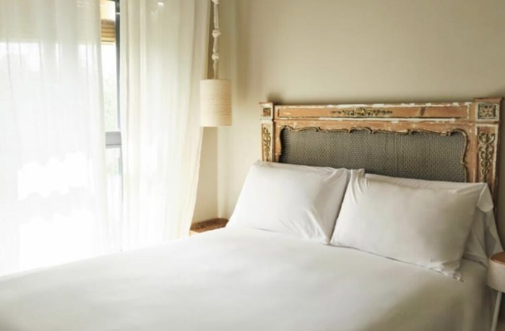 Neatly made double bed with white pillows and a carved antique headboard, beside a window with sheer curtains and soft daylight