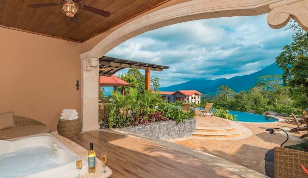 Covered terrace with a corner hot tub and champagne on the rim, looking out to a swimming pool, red-roofed lodges and distant hills