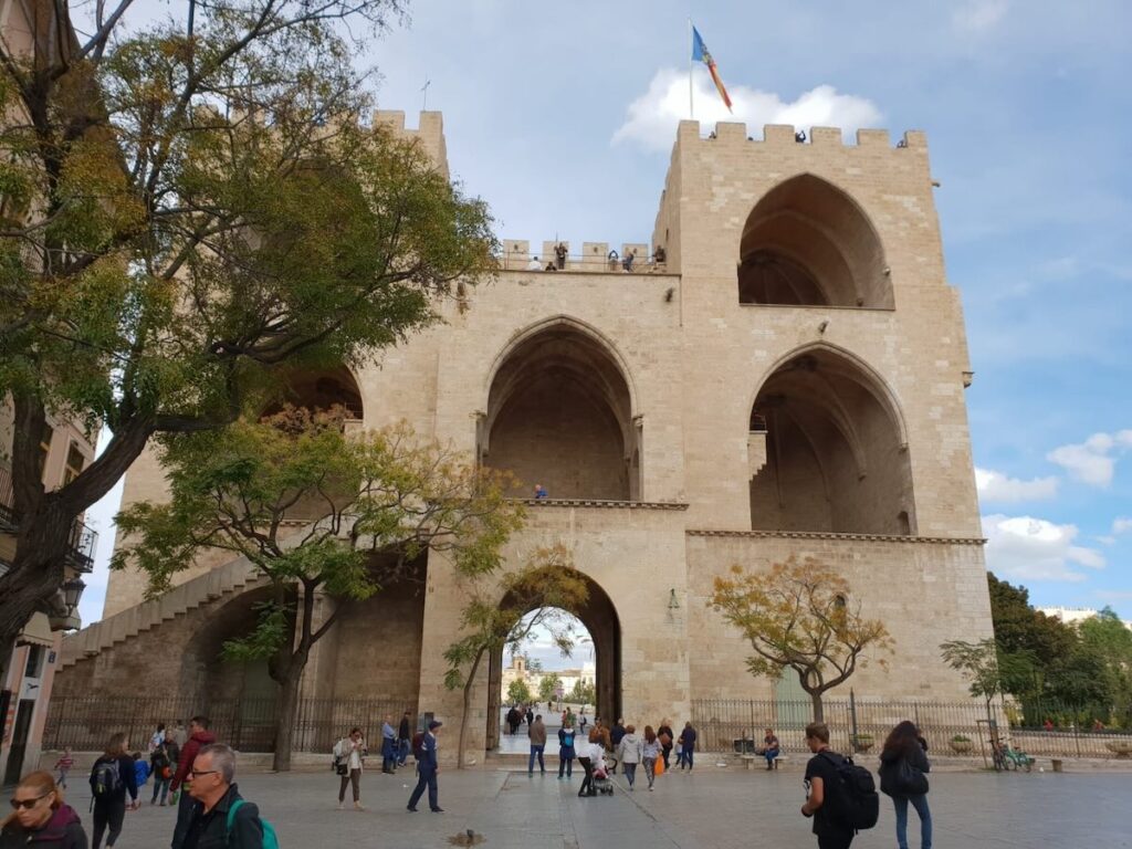 Torres de Serranos, the medieval twin-towered stone gateway in Valencia, with people crossing the tree-lined plaza below