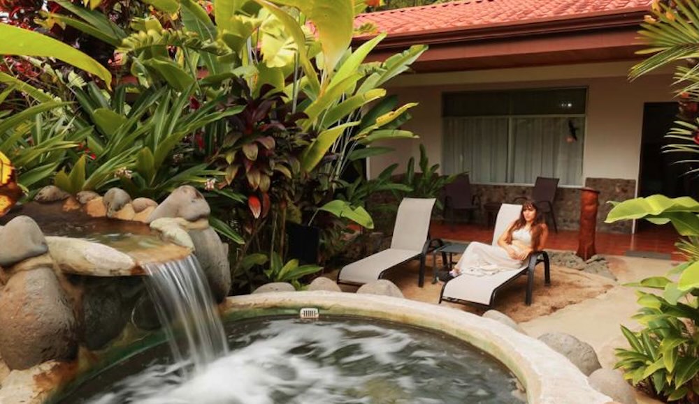 Circular thermal pool fed by a stone waterfall, with two loungers on the sand beside it and a guest room just behind