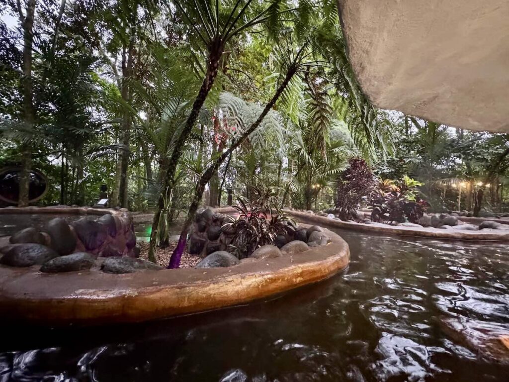 Curving stone thermal hot spring pool surrounded by palms and rainforest at a hotel in La Fortuna, lit at dusk