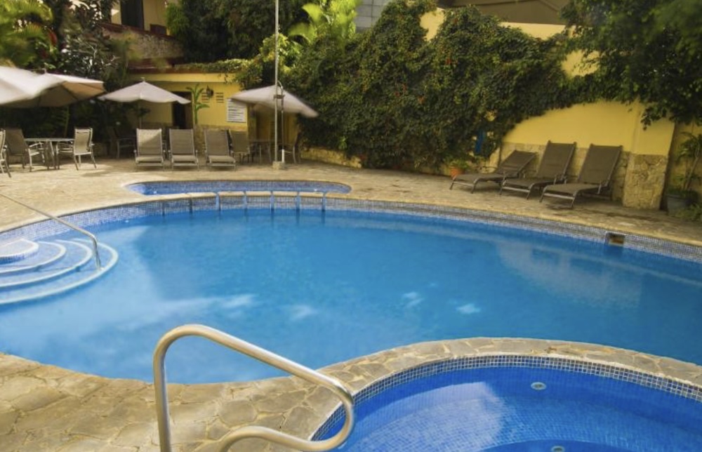 Outdoor pool and separate hot tub edged with blue tiling, sun loungers and parasols on a stone terrace, backed by a wall of climbing greenery