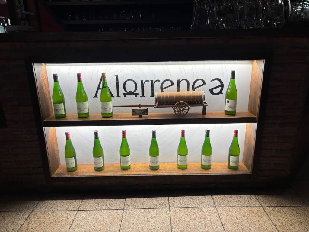 Lit display case of green Alorrenea cider bottles arranged either side of a model wooden barrel cart, with glasses racked above.