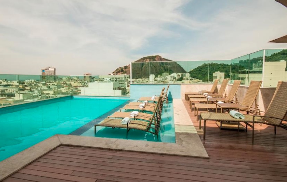 Rooftop pool with a wooden deck and rows of wicker sun loungers, glass safety panels and a rocky green hill rising above the surrounding rooftops