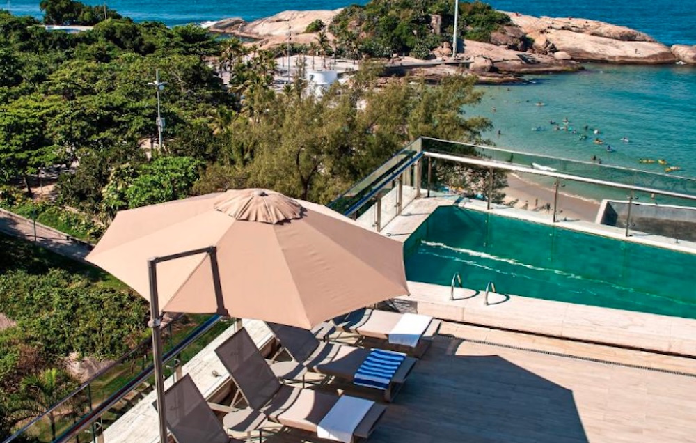 Rooftop pool with a parasol and sun loungers, looking down over trees to the rocky headland at Arpoador and swimmers in the sea beyond