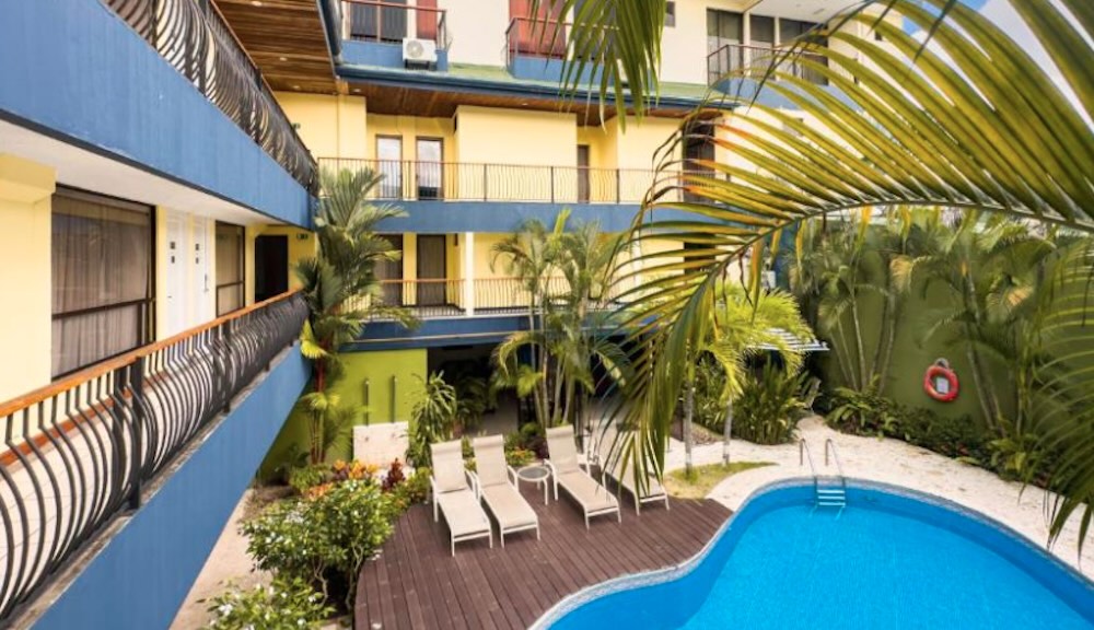 Kidney-shaped pool and wooden sun deck in a courtyard, framed by blue and yellow balconies and tall palms