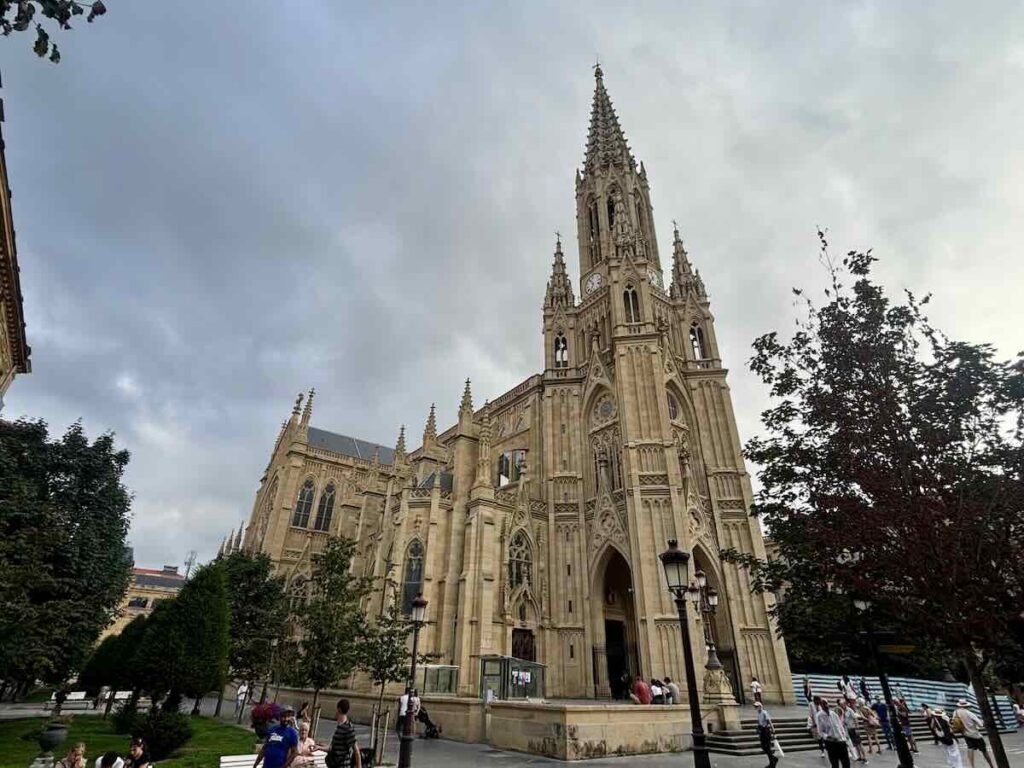 A tall sandstone Gothic cathedral with a central spire, clock face and flanking pinnacled towers, seen from a tree-lined square under grey cloud.