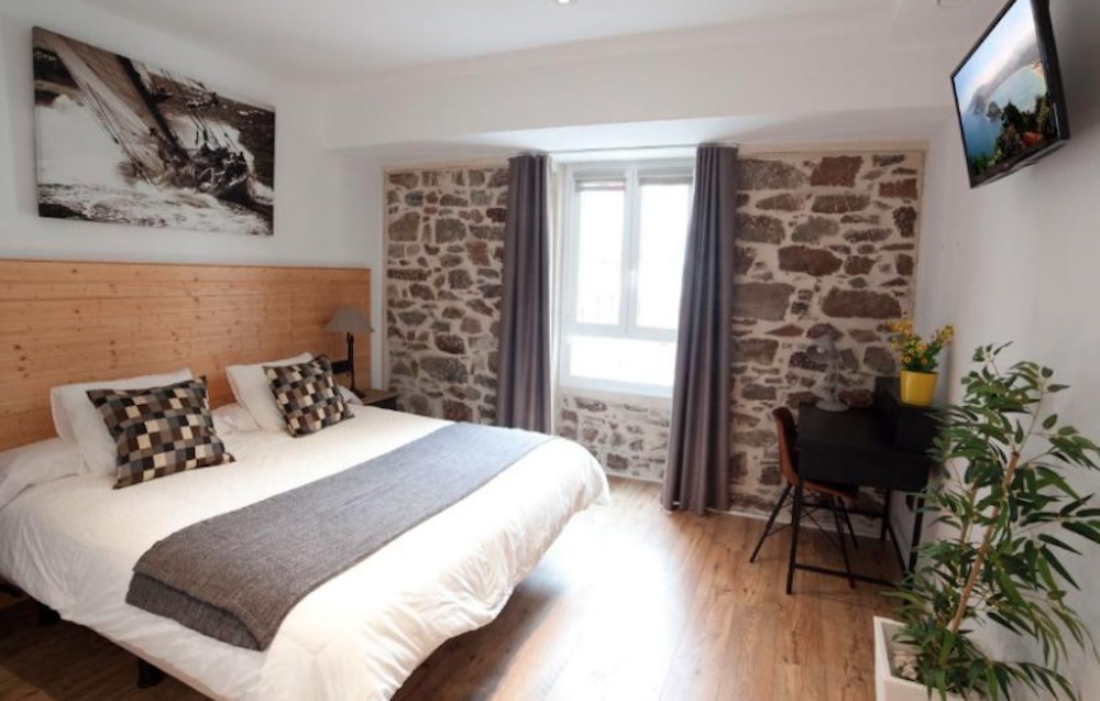 Exposed stone walls framing the window, a wide wooden headboard, grey bed runner, a small black desk and a potted olive plant in the corner.