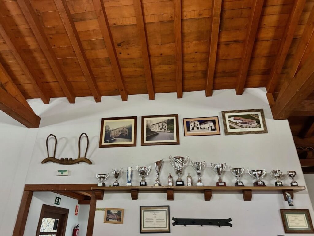 Beamed wooden ceiling above a white wall lined with silver cider trophies and framed photographs of the farmhouse through the years.
