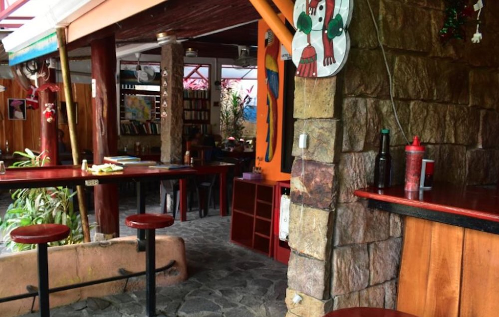 Open-air restaurant with red and orange painted pillars, high stools and a bookshelf along the back wall