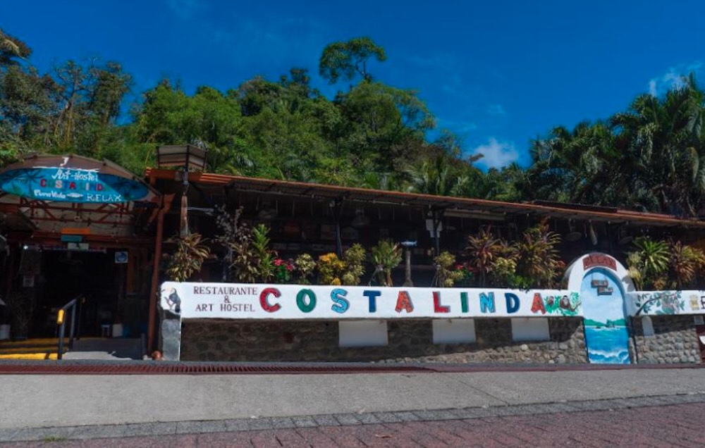 Brightly painted hostel frontage with a hand-lettered sign, backed by dense green rainforest under a blue sky