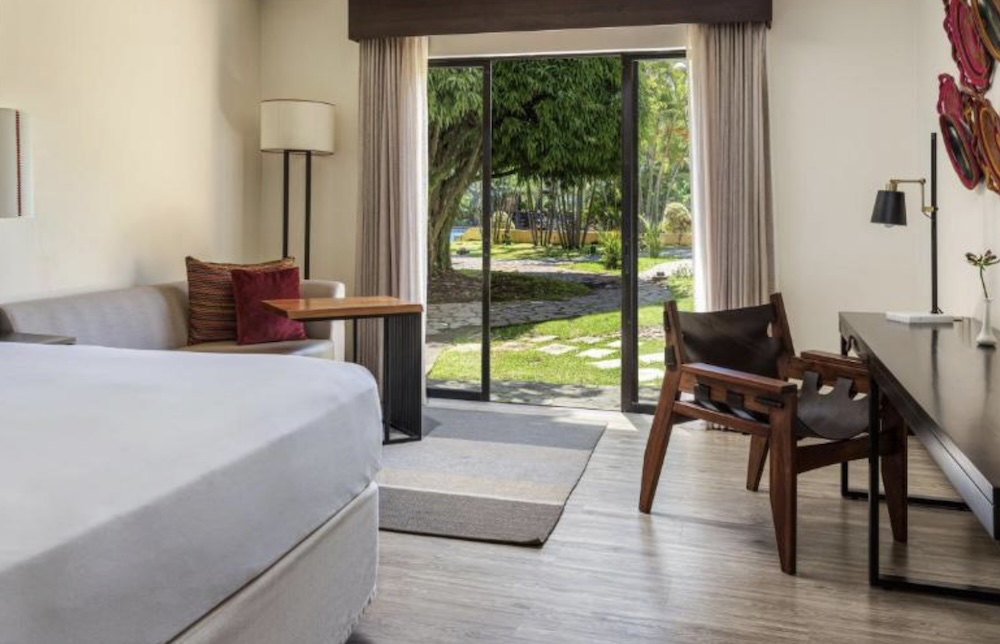 Hotel room with a white bed, wooden desk and leather chair, sliding glass doors opening onto a stone path through a shaded tropical garden