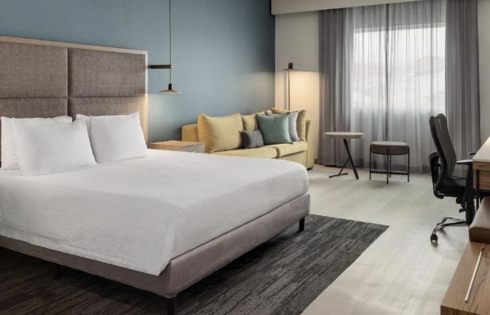Modern hotel room with a king bed, blue feature wall, yellow sofa, desk with an office chair and floor-to-ceiling curtains at the window