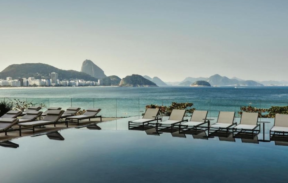 Infinity pool lined with sun loungers, looking out over a curving beach and calm sea towards Sugarloaf Mountain and the hills beyond
