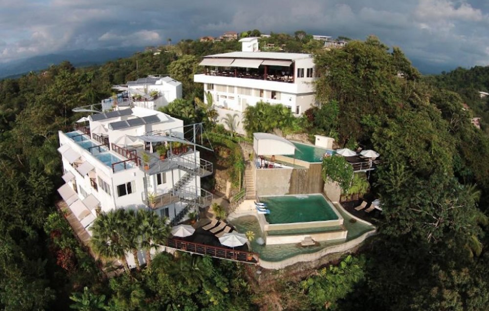 Aerial view of white multi-level buildings and two tiered pools built into a forested hilltop, surrounded by dense jungle