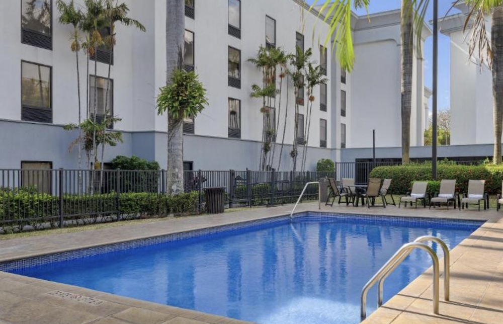 Rectangular outdoor pool with sun loungers and a patio table, enclosed by black railings and palm trees beside a white four-storey hotel