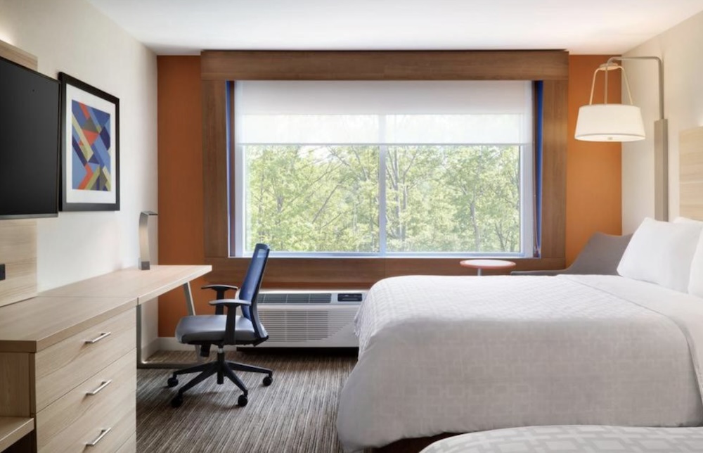 Hotel room with two white beds, an orange feature wall, a long wooden desk with a blue office chair and a large window looking onto trees