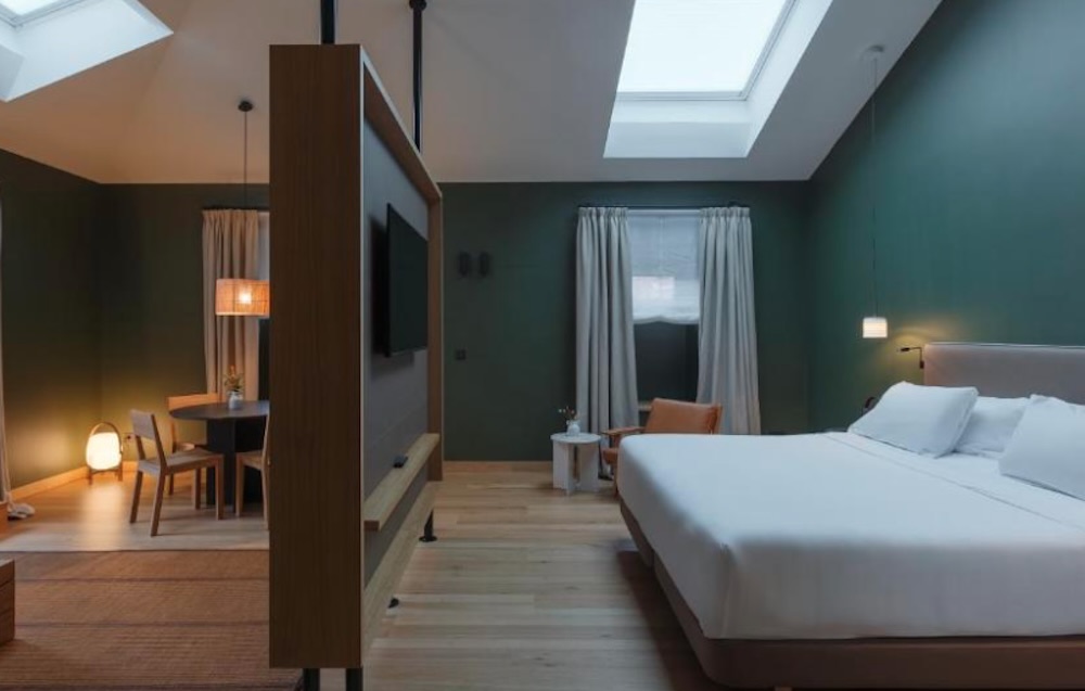 Dark green suite with skylights overhead, a wood partition holding the television, a double bed on one side and a dining table with pendant lights.