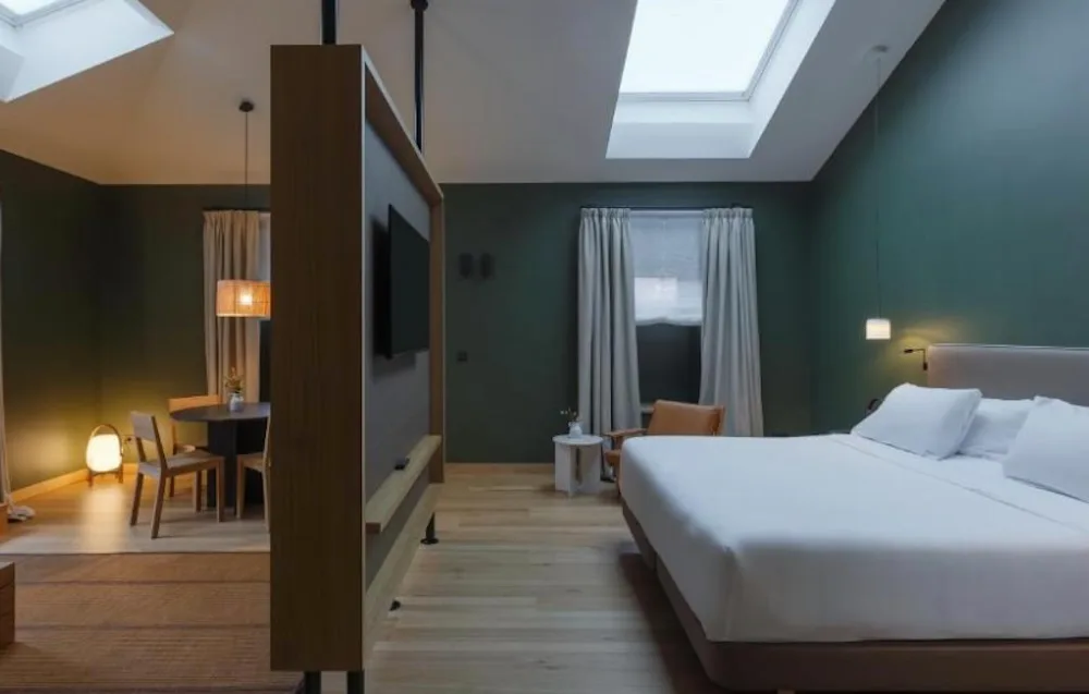 Dark green suite with skylights overhead, a wood partition holding the television, a double bed on one side and a dining table with pendant lights.