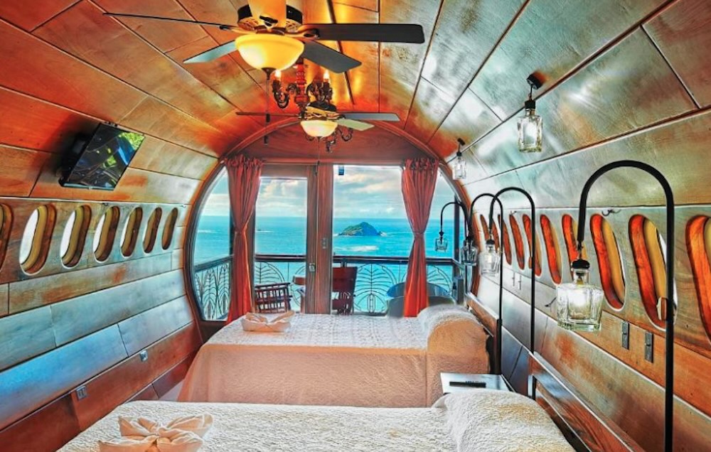 Bedroom inside a converted aeroplane fuselage, with porthole windows, two beds and a balcony overlooking the ocean