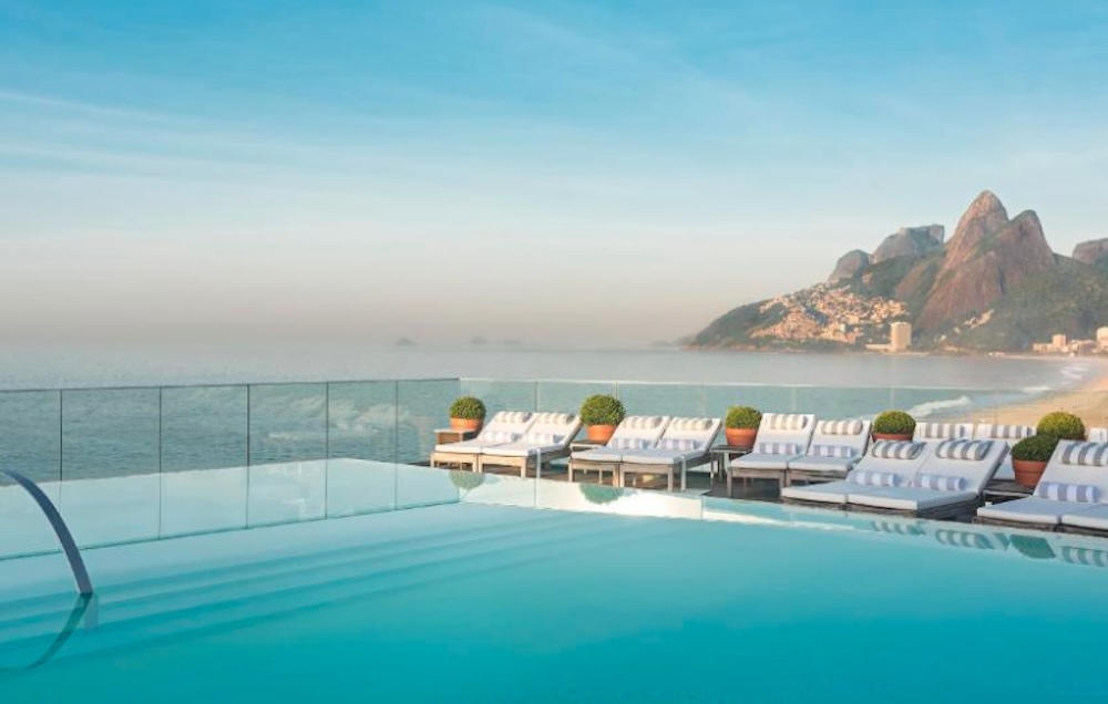 Infinity pool edged with striped sun loungers and clipped topiary, overlooking a wide beach at dawn with Dois Irmãos rising at the far end