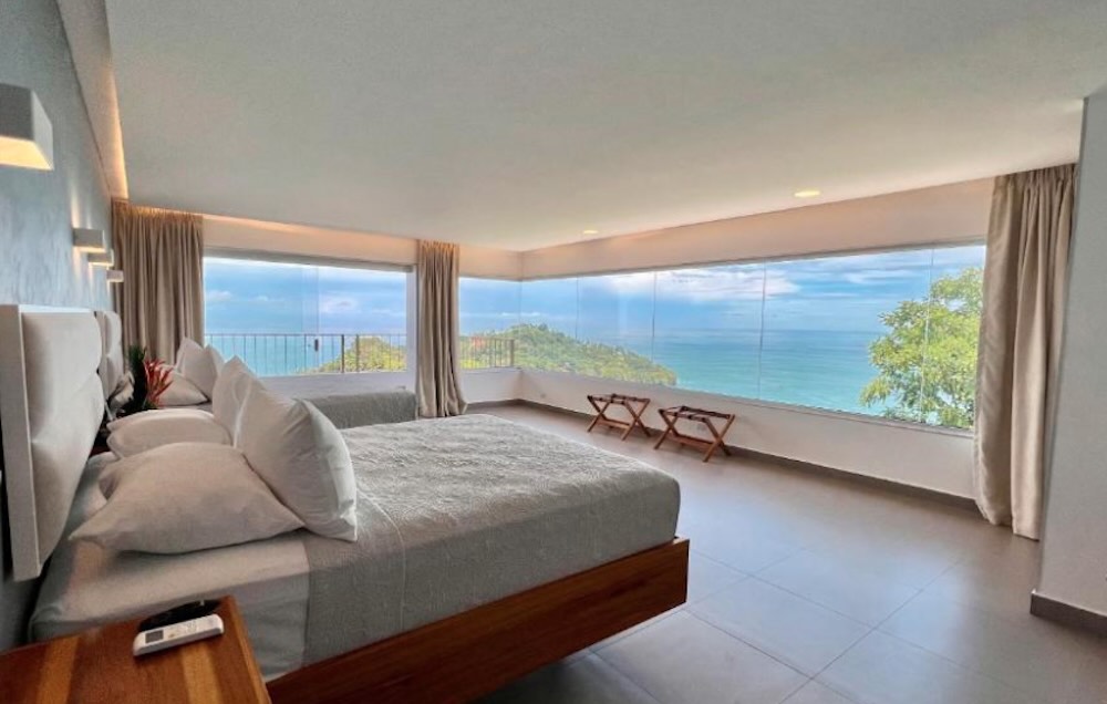 Minimal bedroom with a low wooden bed and wraparound picture windows framing a headland and the ocean below
