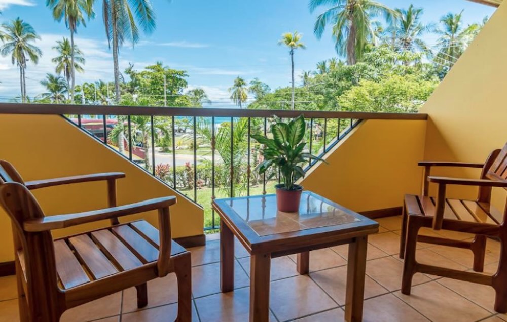 Balcony with wooden chairs and a table, looking past palm trees to the sea across the road