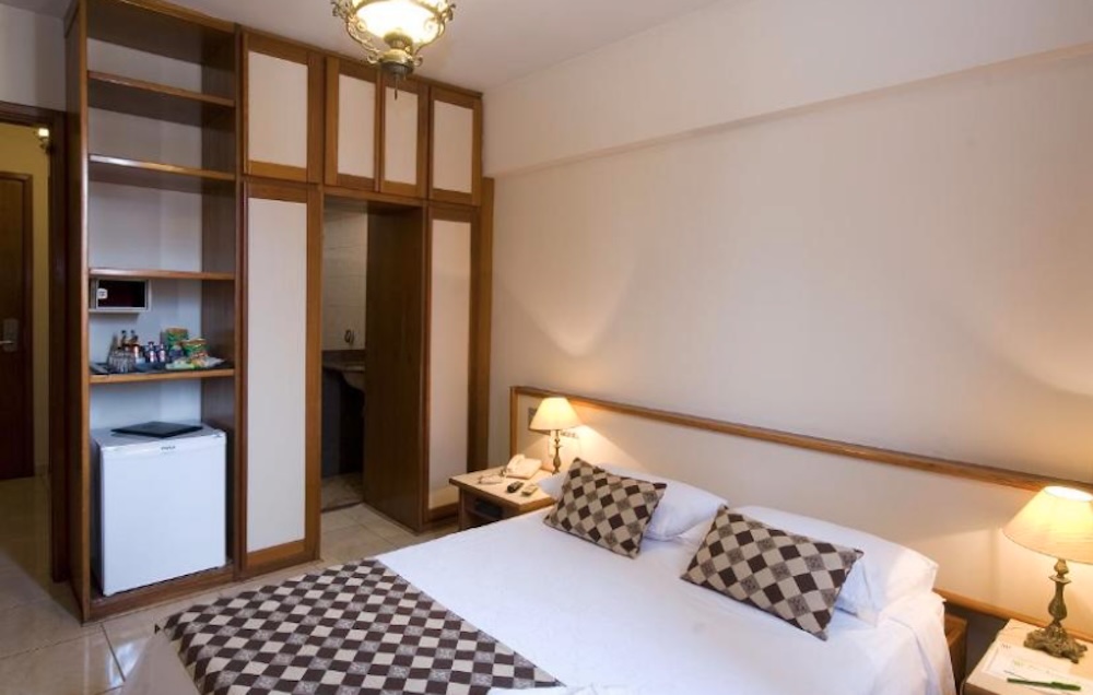 Simple double room with a fitted dark wood wardrobe, open minibar shelf with a small fridge, bedside lamps and a brown checked bed runner