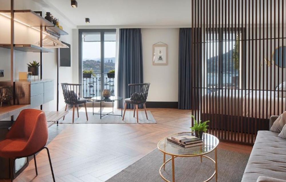 Open plan suite with a slatted wood screen dividing the bedroom, a sofa and low round table, and glazed doors onto a balcony above the harbour.