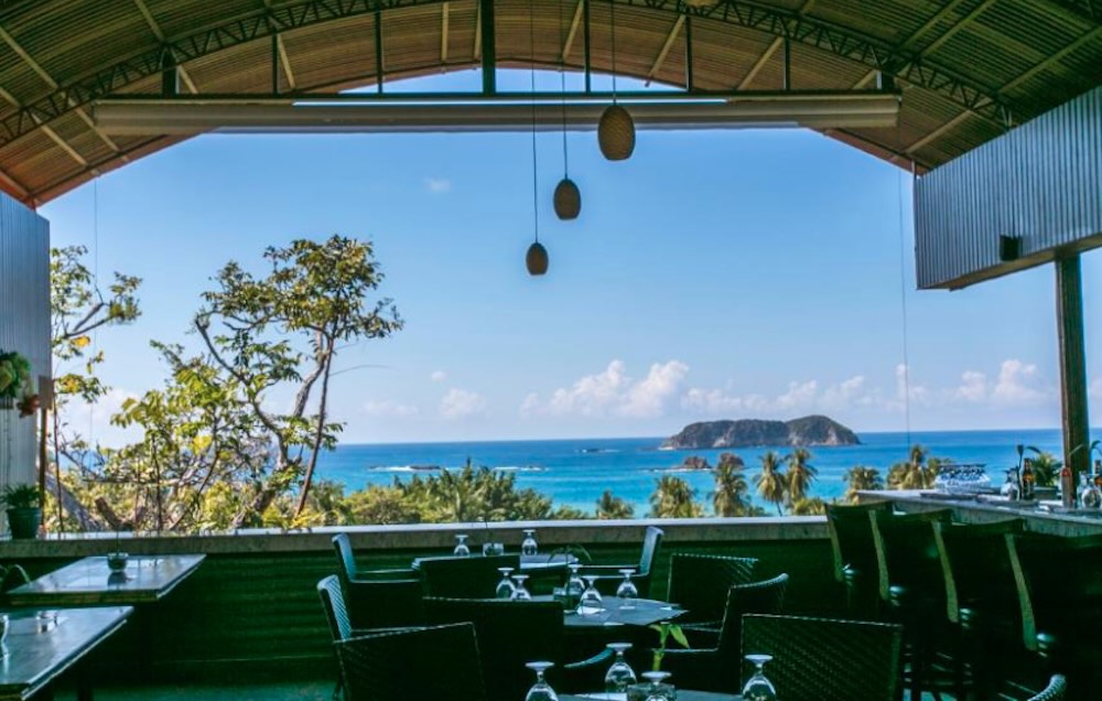 Open-sided hilltop restaurant with laid tables, looking out over treetops to the ocean and a rocky island