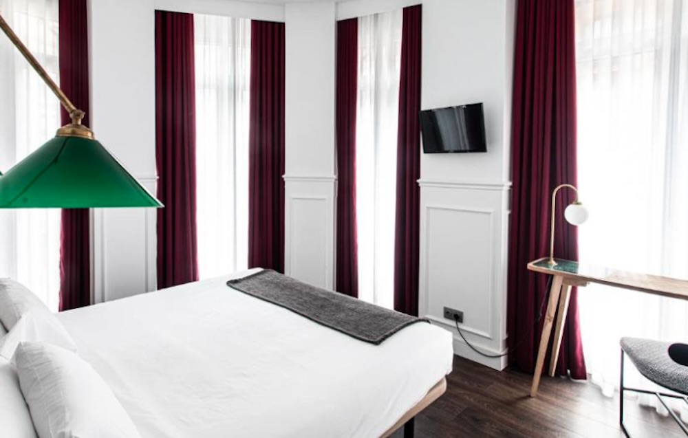 White panelled bedroom with three tall windows in deep red curtains, a green glass reading lamp above the bed and a slim desk on dark wood flooring.