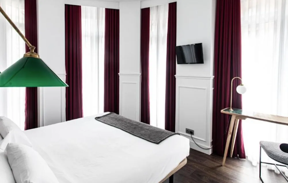 White panelled bedroom with three tall windows in deep red curtains, a green glass reading lamp above the bed and a slim desk on dark wood flooring.