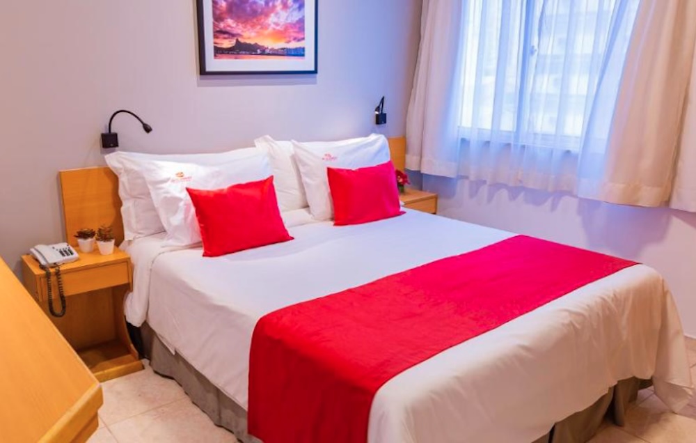 Simple double room with white bedding, red cushions and a red bed runner, pale wood bedside tables and a framed photograph of a city sunset above