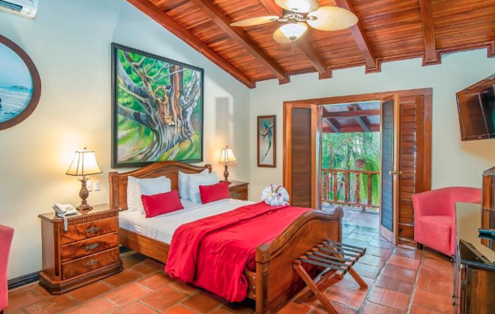 Bedroom with a dark wooden beamed ceiling, terracotta floor, red bedspread and open doors onto a balcony