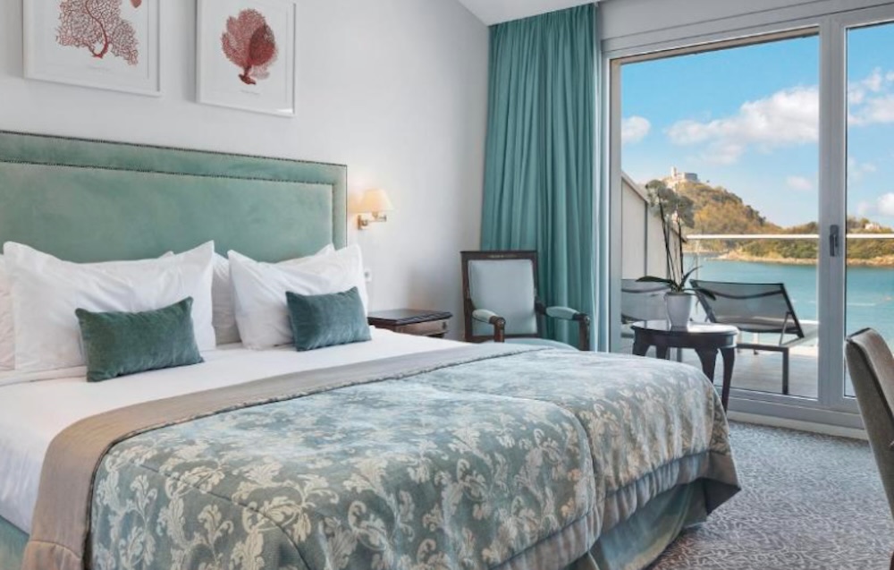 Green upholstered headboard and patterned bedspread facing open balcony doors, with a wooded headland and still bay filling the view beyond.