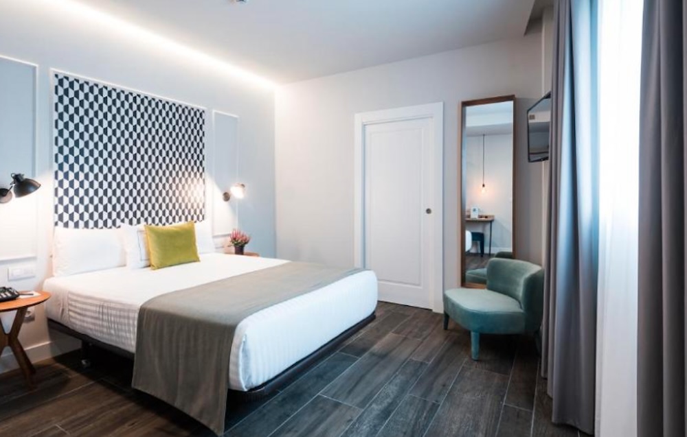 Pale blue bedroom with a black and white chequered headboard panel, a yellow cushion on the bed, a teal armchair and a tall mirror by the doorway.